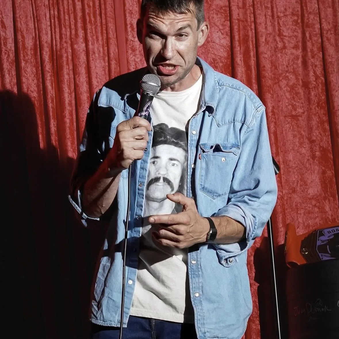 Shane Camp performing in front of red curtains wearing a Glen Wayne Albert shirt