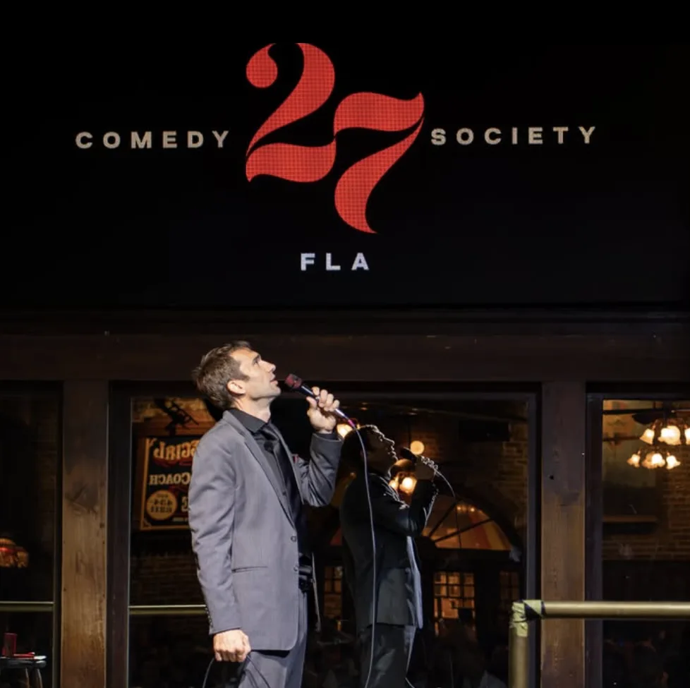 Shane Camp performing onstage beneath a Comedy 27 Society sign